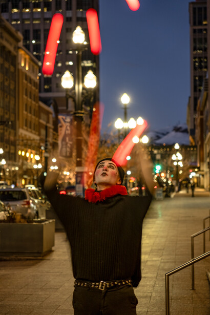 Person in red beanie and black jacket with arms raised toward illuminated clock tower at dusk on urban street.