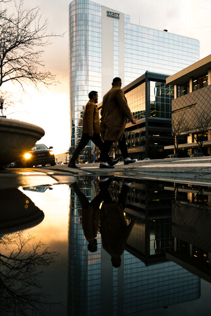 Two figures walking past a modern glass building reflected in a wet pavement puddle at golden hour.