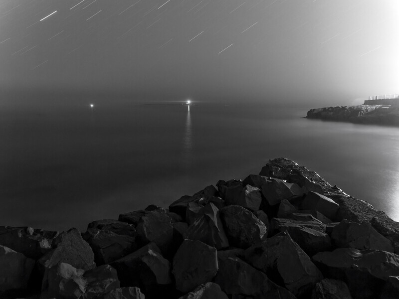 Rocky breakwater in foreground with calm water and distant pier lights under overcast night sky.