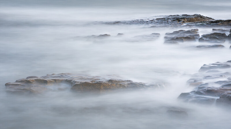 Misty seascape with layered rocky outcrops partially submerged in soft, flowing water.