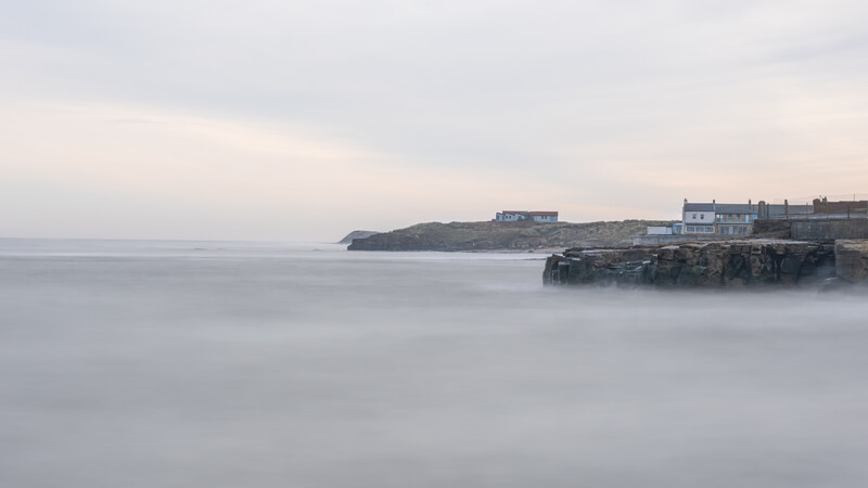 Coastal houses perched on rocky cliffs overlooking misty seawater at dusk.