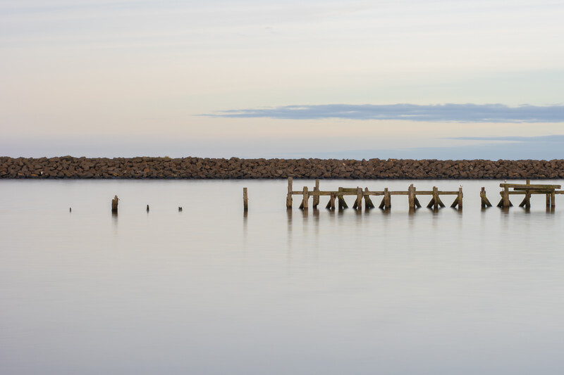 Wooden pilings and a deteriorating dock structure reflected in still water at dusk.