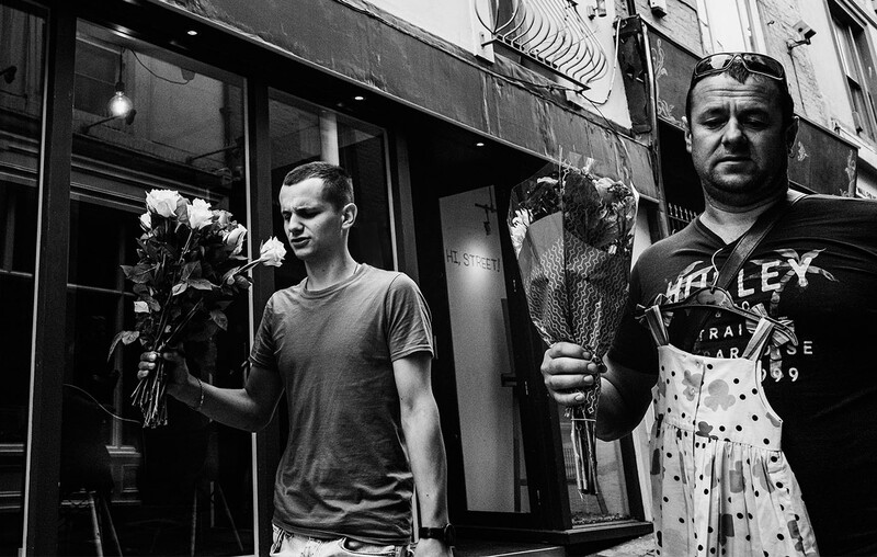 Two men holding wilted flowers and dried plants on an urban street in black and white.
