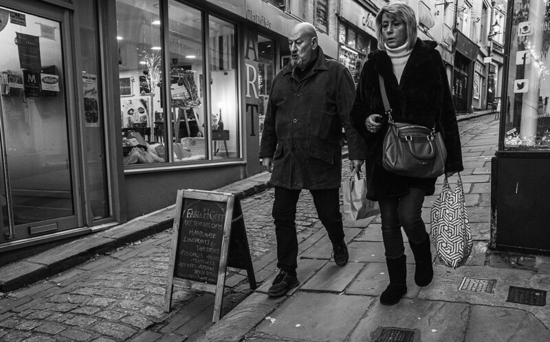 Two people walking past storefronts on a cobblestone street in black and white.