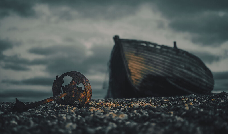 Rusted metal ring on pebbled beach with abandoned wooden boat in background under stormy sky.