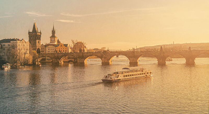 Passenger riverboat on a waterway beneath an arched stone bridge with Gothic architecture and towers along the embankment.