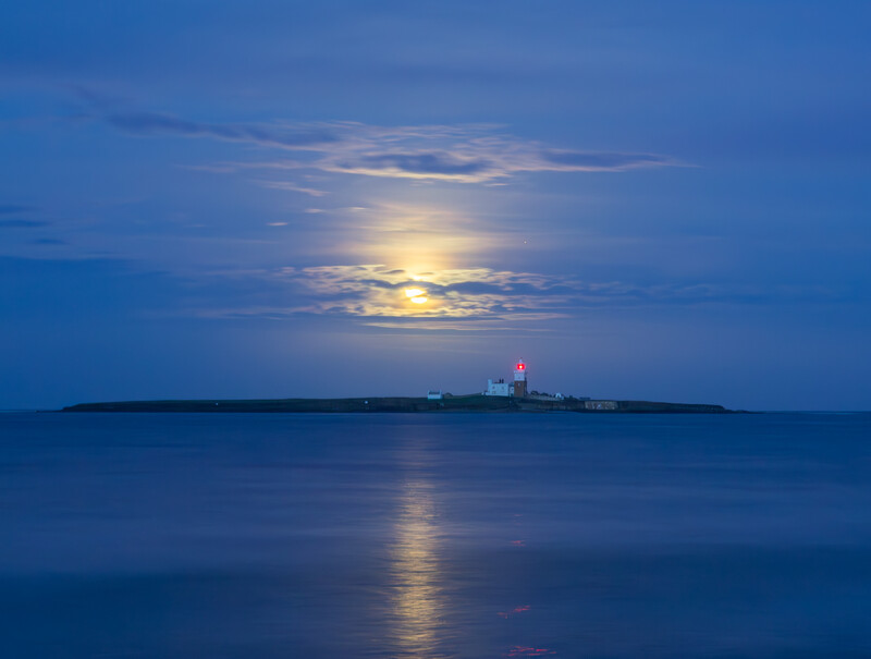 Lighthouse on a distant island during twilight, with moonlight reflecting across calm water.