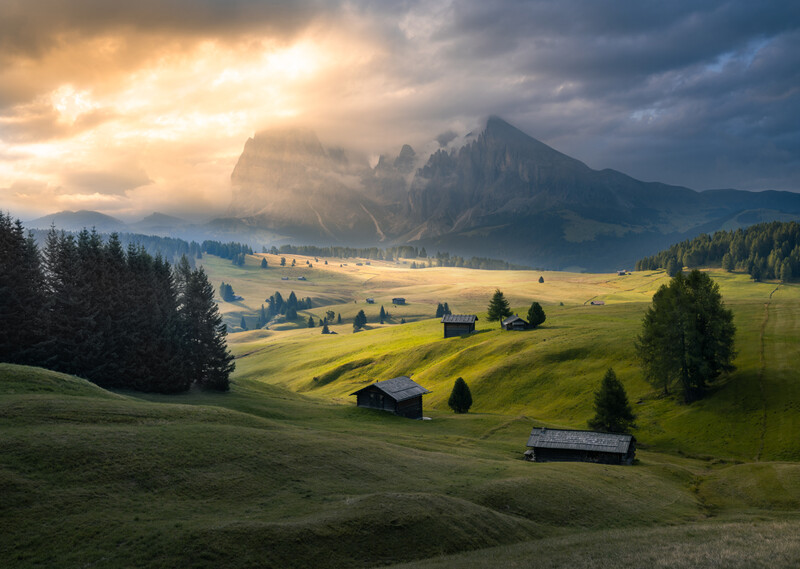Alpine valley with scattered farmhouses, dramatic mountain peak, and golden light breaking through storm clouds.
