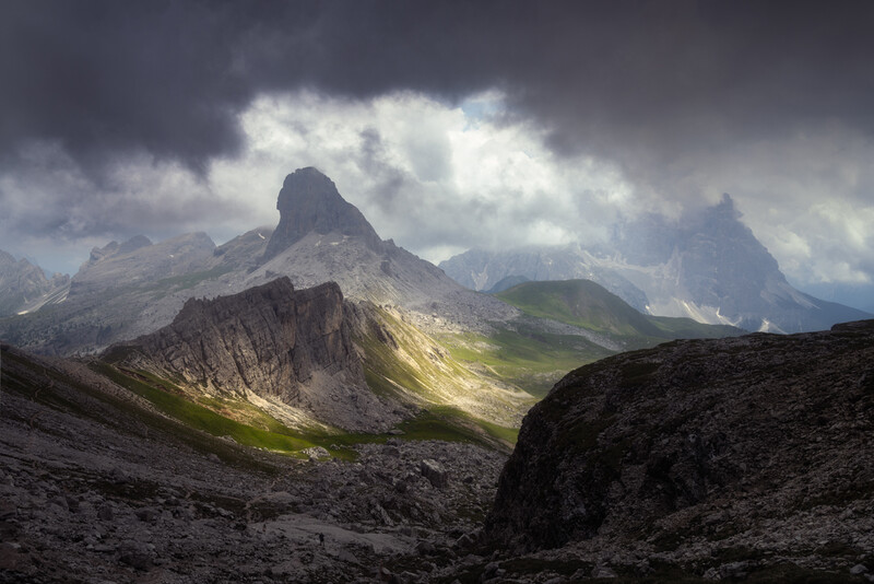 Alpine mountains with dramatic storm clouds and a sunlit valley between dark rocky foreground peaks.