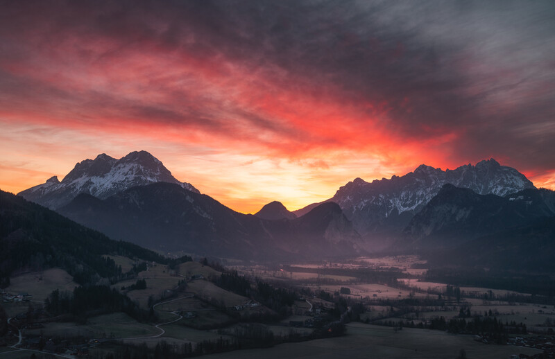 Snow-capped mountain peaks silhouetted against a dramatic crimson and gold sunset sky.