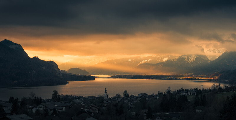Alpine lake at sunset framed by dramatic mountain peaks and storm clouds with golden light breaking through.