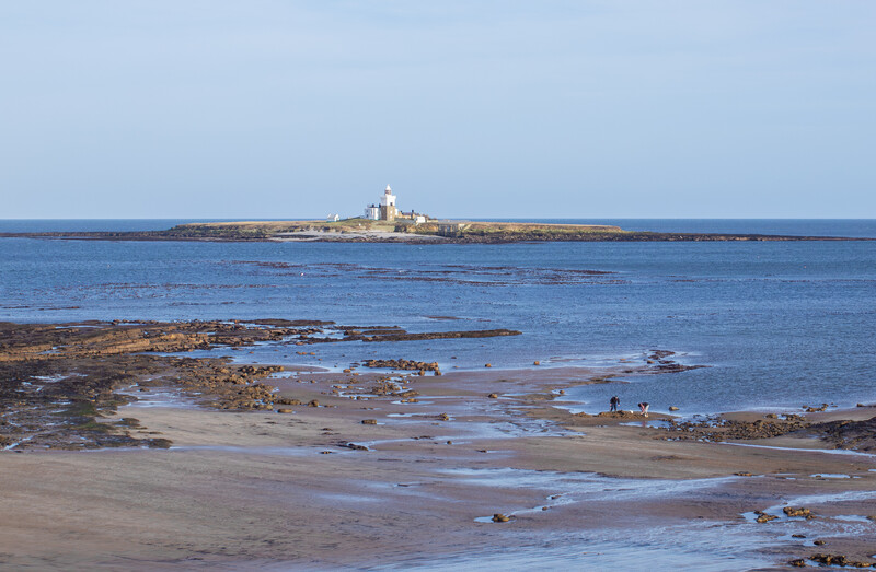 Rocky coastline with tidal pools and a distant lighthouse on the horizon under clear blue sky.