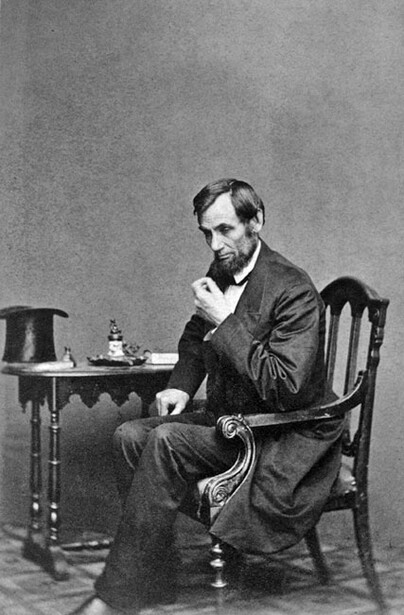 Bearded man in dark suit seated at small table, resting chin on hand in contemplative pose.