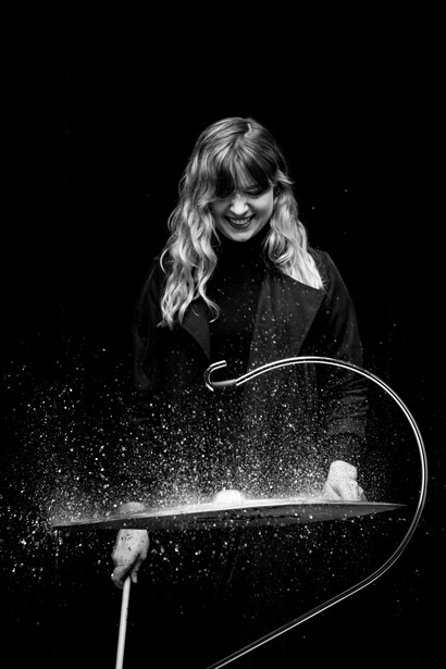 Woman with long blonde hair playing electric violin against black background with light trails.
