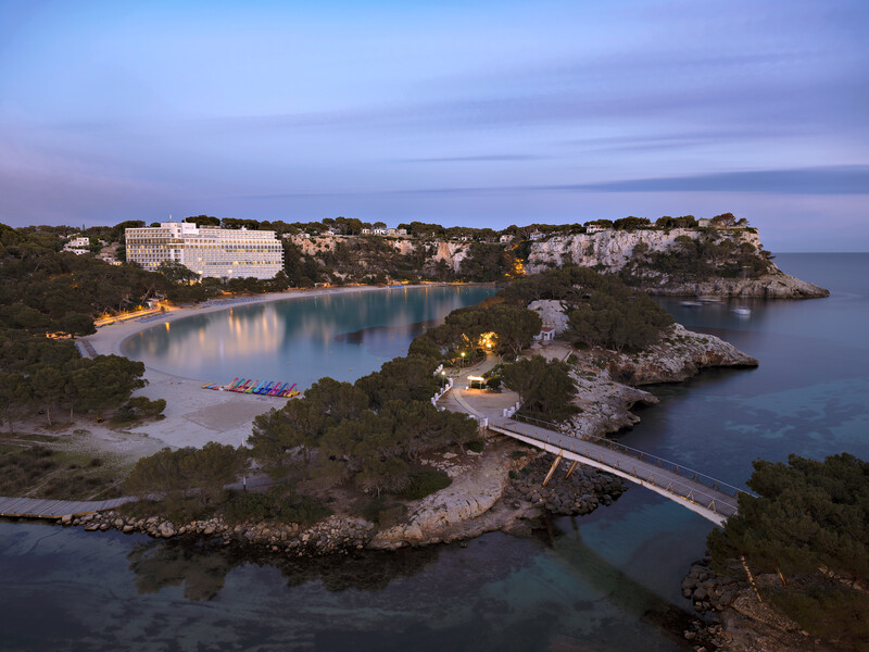 Coastal cove at twilight with illuminated resort hotel, forested cliffs, and calm turquoise water.