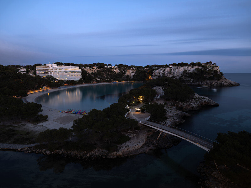 Coastal resort at dusk with illuminated buildings overlooking a curved bay surrounded by rocky peninsulas.