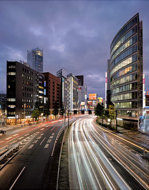 Urban street at dusk with light trails from vehicle traffic flowing through a multi-lane road flanked by illuminated buildings.