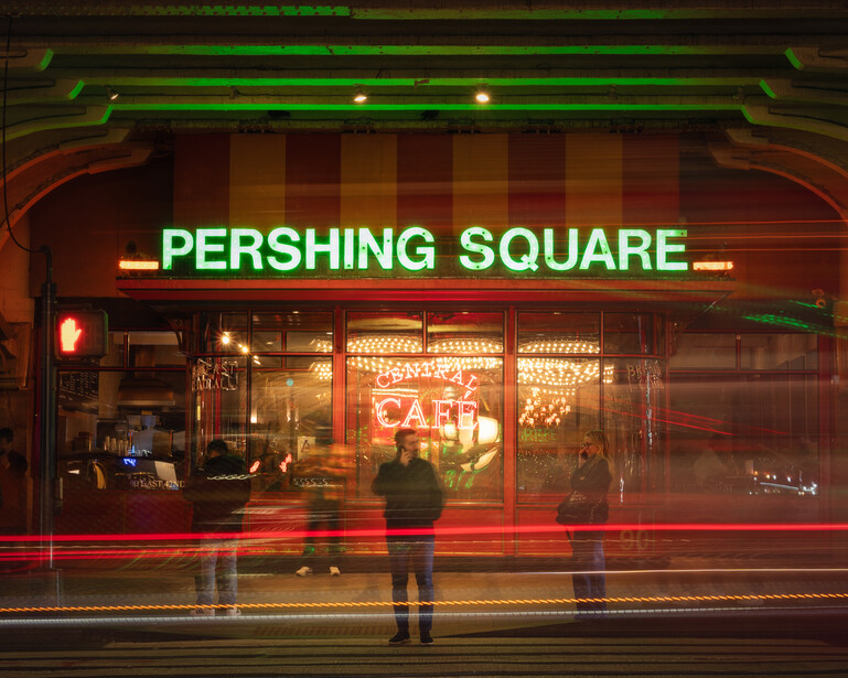 Neon storefront with green sign reading "PERSHING SQUARE" above shop windows lit in warm amber tones, with blurred figures on street at night.