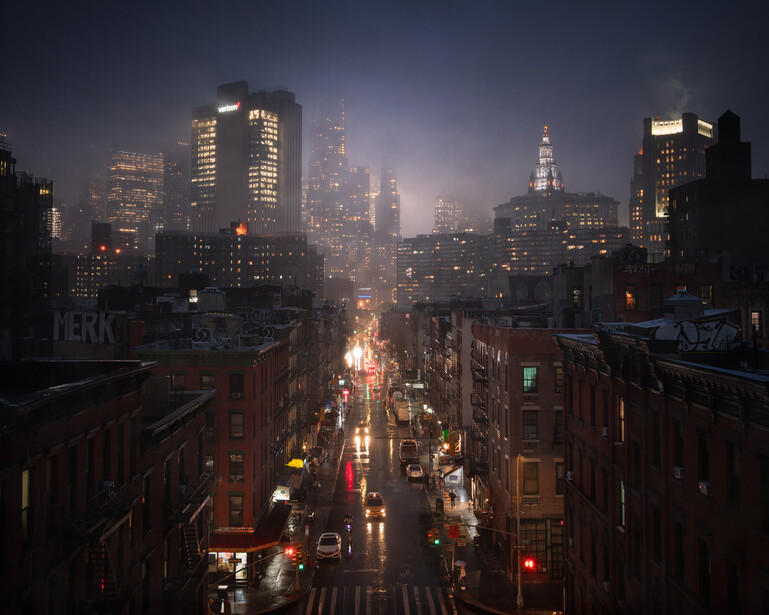 Cityscape at dusk showing a street-level view between tall buildings with illuminated windows and street lights.