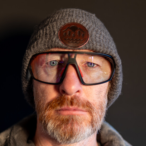 Man wearing glasses and gray beanie with patch, studio portrait with neutral background.