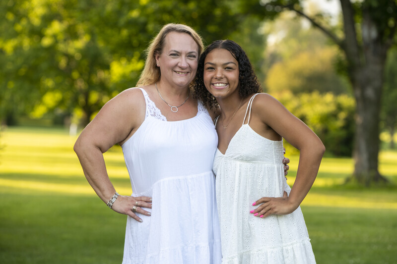 Two women in white dresses pose together outdoors in natural daylight.