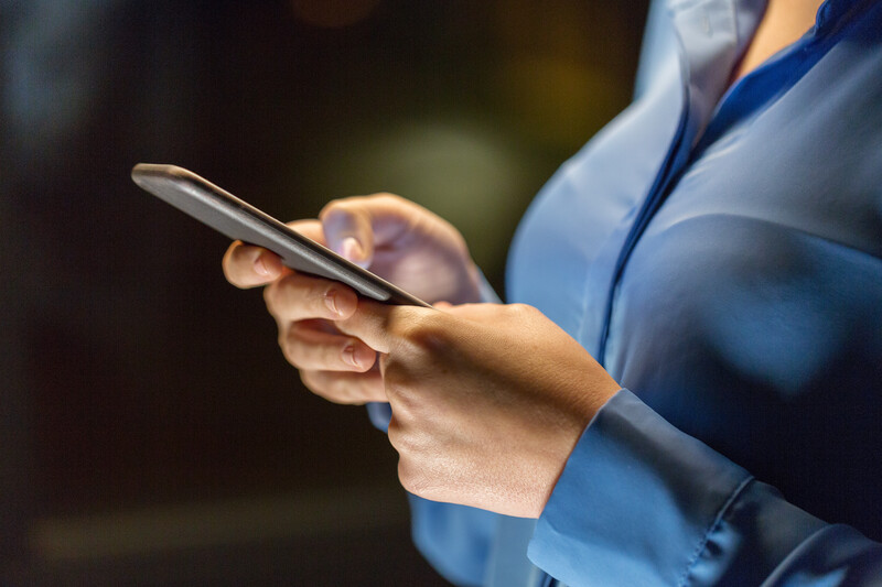 Person in blue shirt holding smartphone with both hands, photographed from chest up with blurred background.