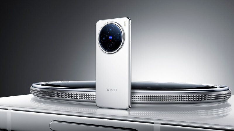 White smartphone with circular camera module displayed upright against curved metallic reflective surfaces.