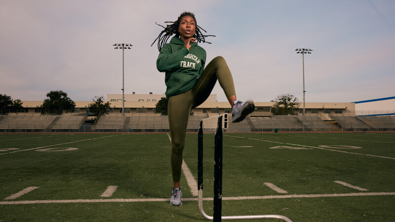 Athlete in green tracksuit high-kicking a pole on a stadium field at golden hour.