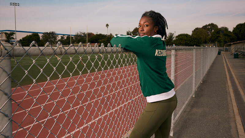 Athlete in green team jersey posing at chain-link fence bordering a red clay tennis court.