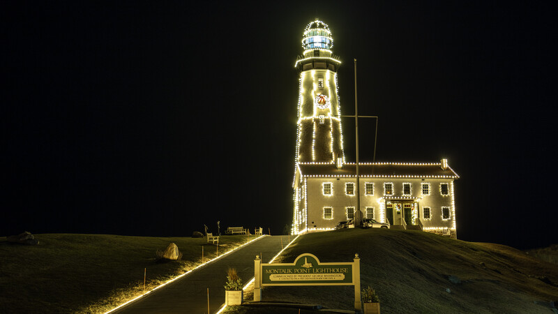 Illuminated lighthouse on a hill at night with warm golden outline lighting and adjacent keeper's house.