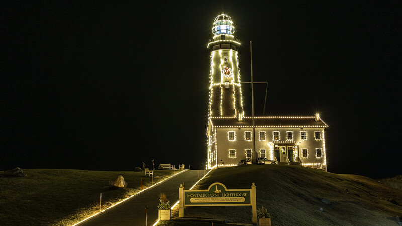 Illuminated lighthouse on a sand dune at night with attached keeper's house glowing in warm golden light against dark sky.