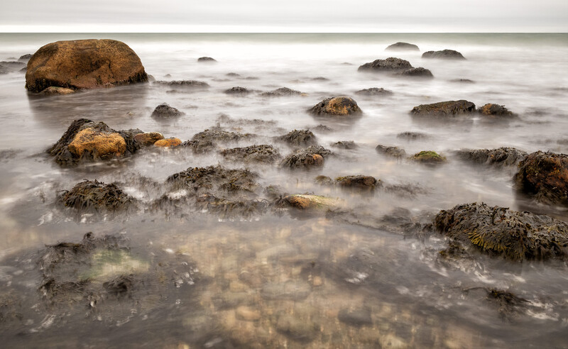 Rocky coastline with long-exposure blurred water and scattered boulders at surf line.