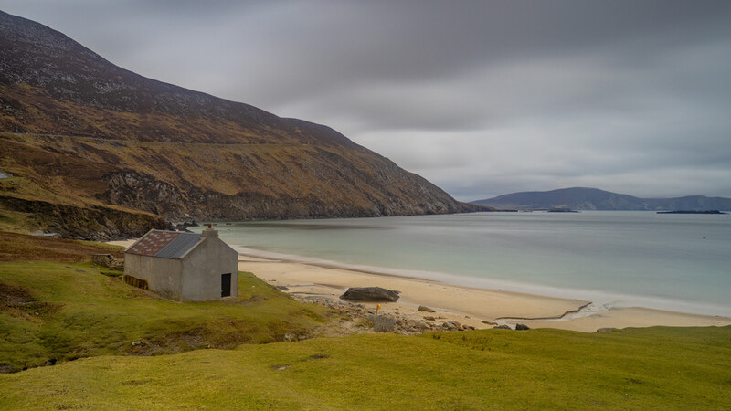 Abandoned cottage on grassy slope overlooking a turquoise beach and calm sea beneath dramatic clouds.