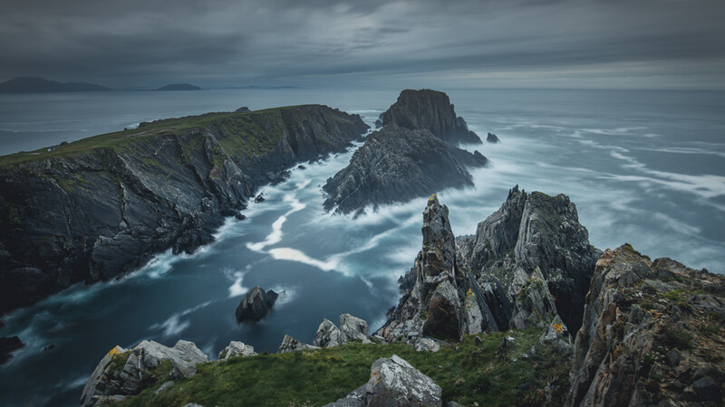 Dramatic rocky coastline with jagged sea stacks and waves crashing against dark cliffs under stormy skies.