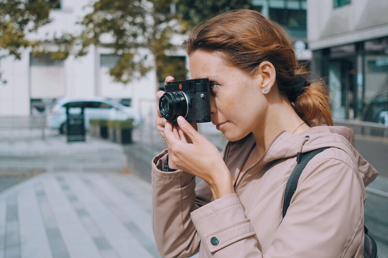 Woman holding a vintage film camera to her face while standing in an urban plaza.