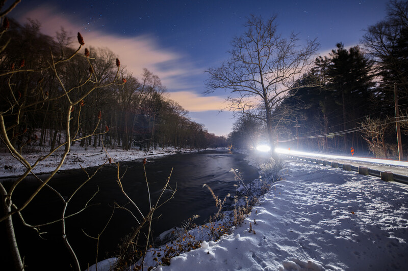 Frozen river at twilight flanked by bare trees with a bright vehicle light source illuminating the snowy landscape.