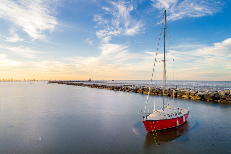 Red and white sailboat moored in calm shallow water with a breakwater visible in the distance under blue sky.