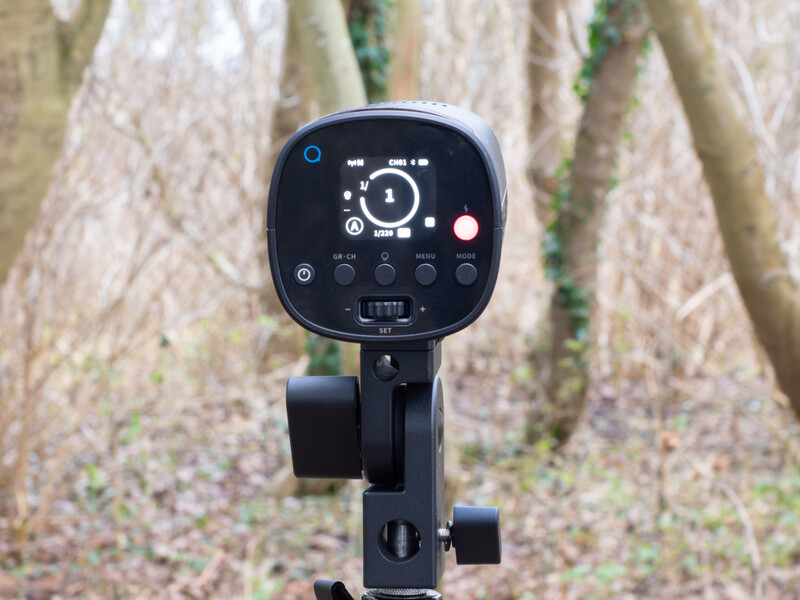 Black compact flash unit with control panel mounted on tripod stand against tree bark background.