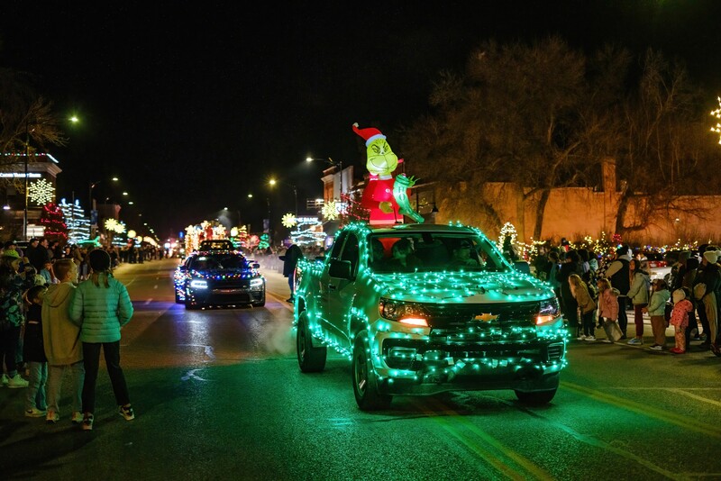 Green LED-decorated truck in a nighttime holiday parade with crowds lining the street.