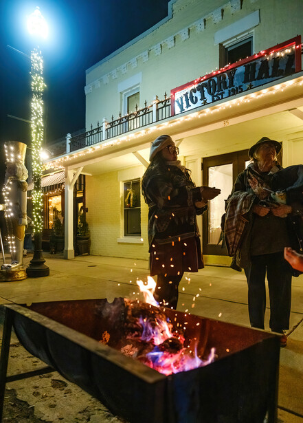 Two people standing near a fire pit at night in front of an illuminated building with holiday decorations.
