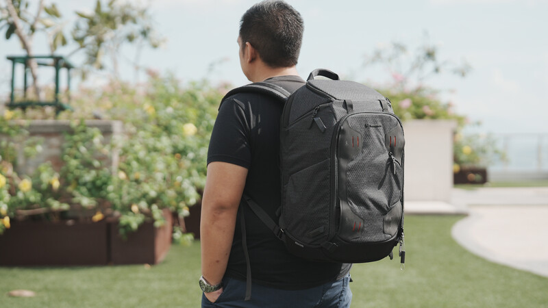 Man wearing a dark gray camera backpack stands in a sunlit rooftop garden with plants and city views.