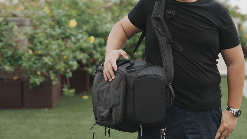 Person wearing black camera backpack over black t-shirt in outdoor garden setting.
