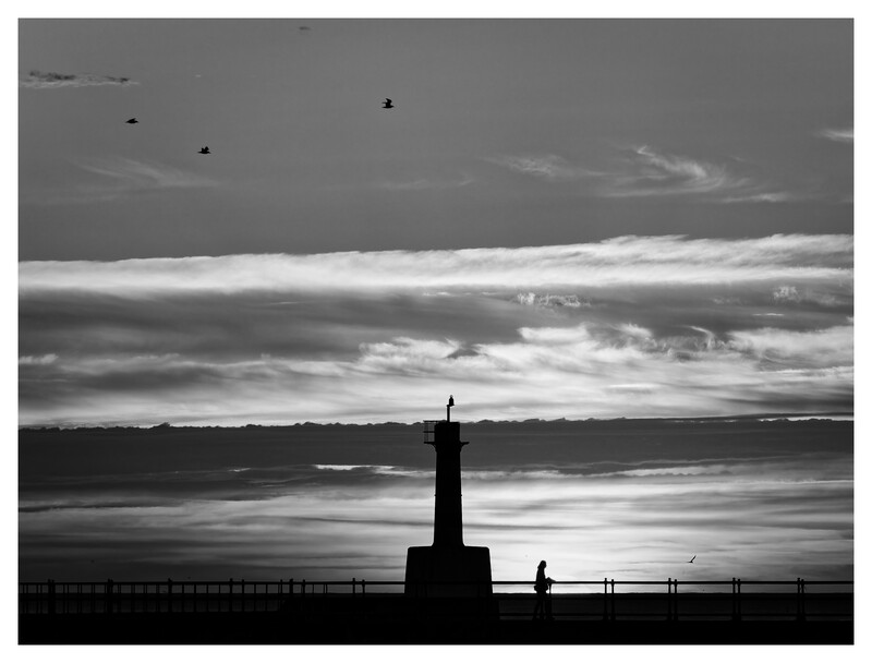 Silhouetted lighthouse and figure on a pier against a dramatic cloudy sky at dusk.