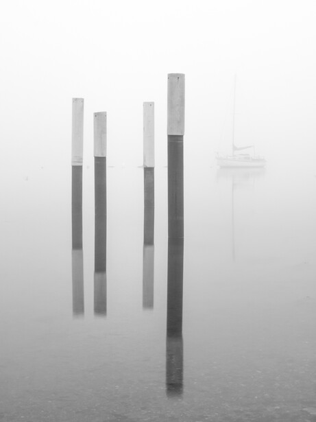 Wooden posts emerging from fog and still water, with a distant structure barely visible through the mist.