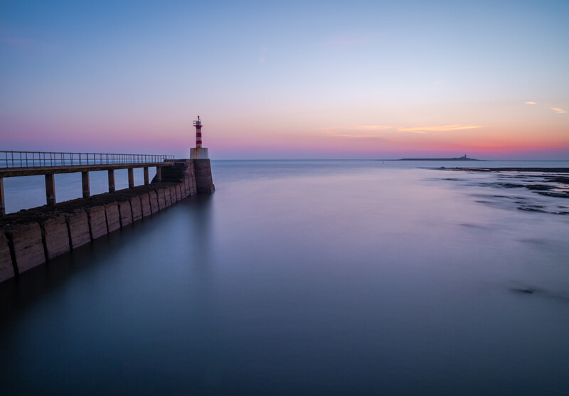 Lighthouse at the end of a long pier extending into calm water at dusk, with pink and purple sky.