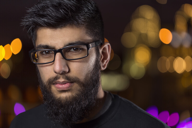 Man with dark beard wearing glasses and black shirt against blurred golden and purple bokeh background.