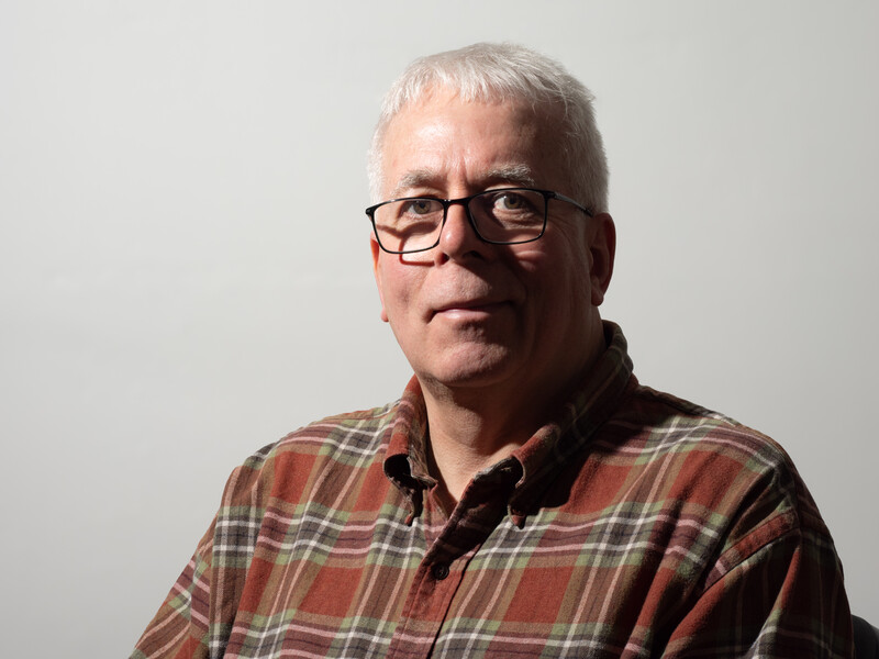 Older man wearing glasses and a plaid shirt against a neutral background.