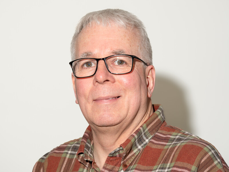 Middle-aged man with gray hair and glasses wearing a plaid shirt, photographed against a white background.
