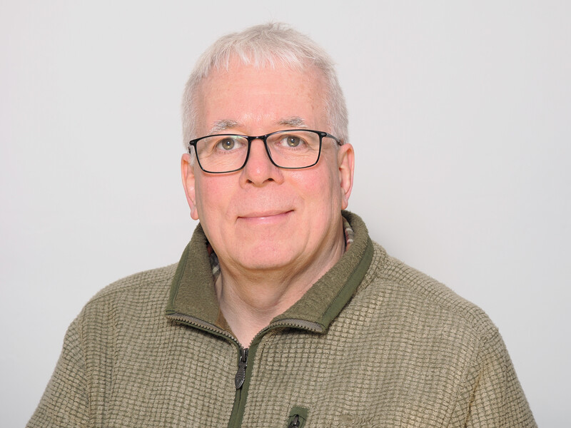 Middle-aged man with white hair and glasses wearing a tan and olive quarter-zip pullover against a plain gray background.