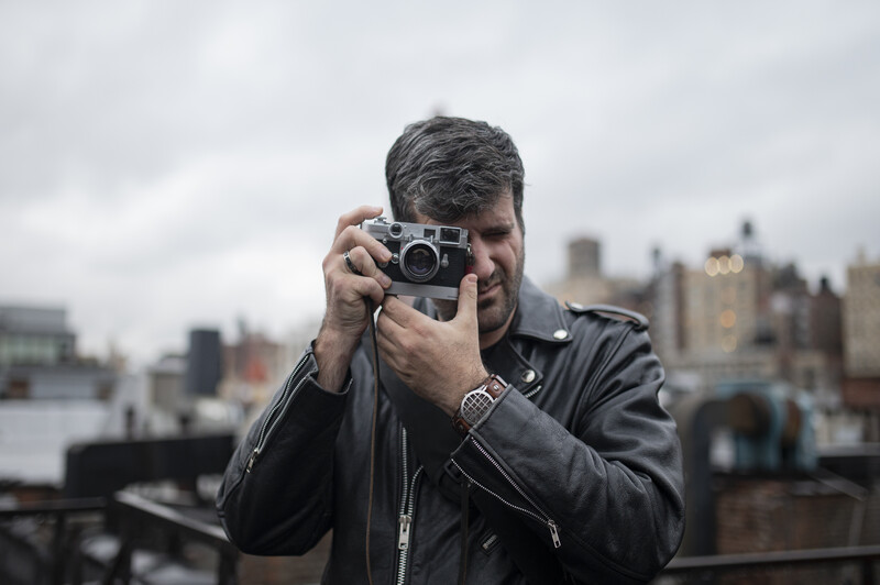 Photographer holding a rangefinder camera up to his face, shooting against an urban skyline.